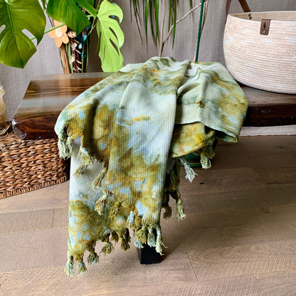 A hand-dyed green cotton throw blanket from Astilbe Lane, shown draped across a wood bench.