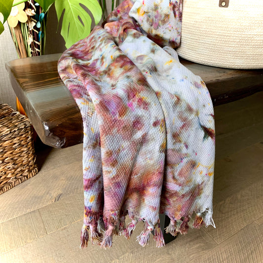 A hand-dyed purple cotton throw blanket from Astilbe Lane, shown draped across a wood bench.