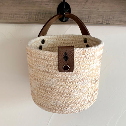 Cream colored rope basket with brown leather strap from Astilbe Lane, shown hanging from a wall hook.